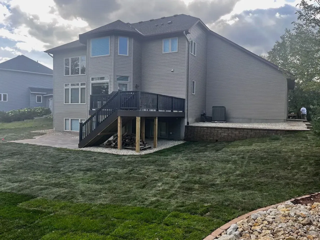 Back of a two-story gray house with a wooden deck and a brick patio. Green lawn slopes towards the edge of the yard.