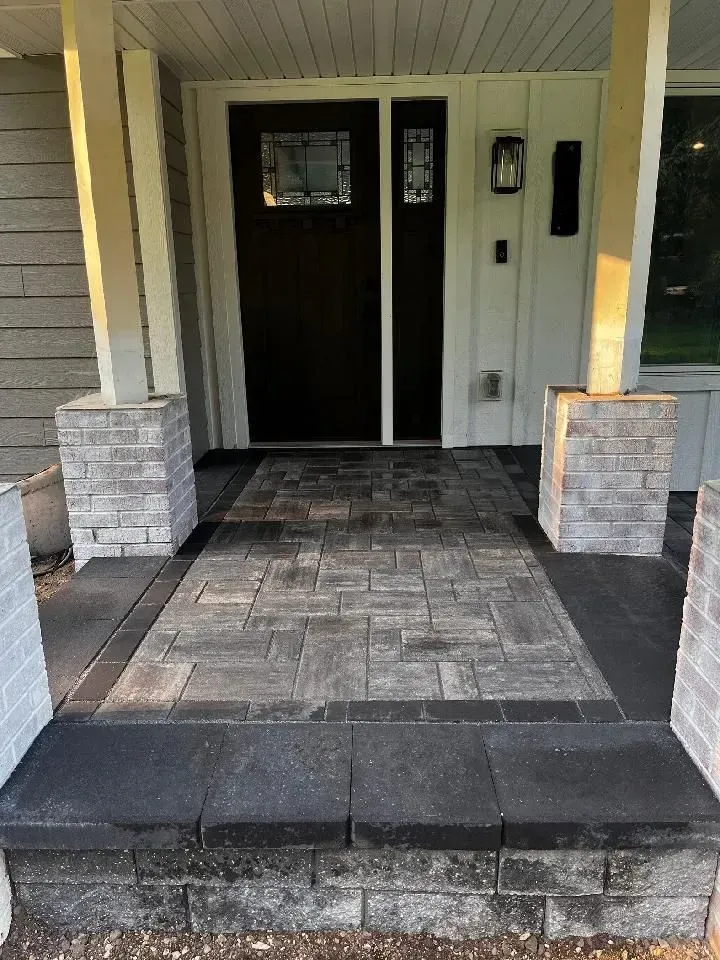 A small patio with curved edges, constructed of light brown pavers, is adjacent to wooden stairs.