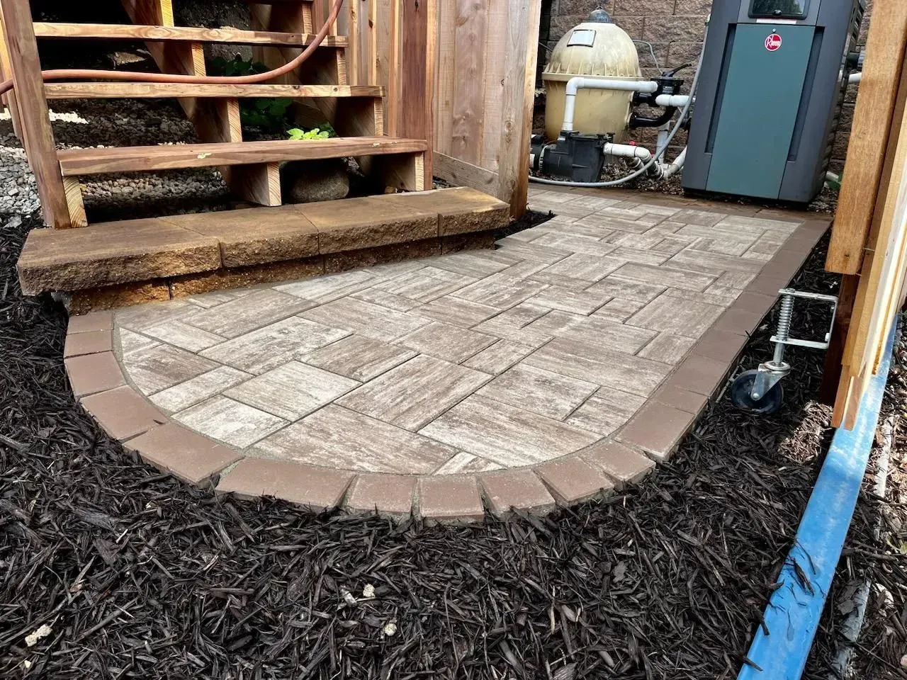 A small brick patio extends from wooden steps. Brown pavers and edging sit on dark mulch a pool pump & equipment are visible.
