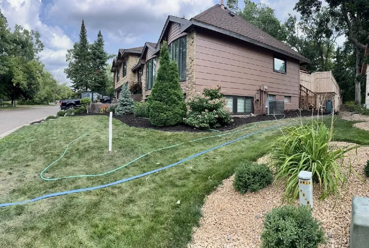 A two-story brown house with lush green landscaping and an irrigation system, on a sunny day.