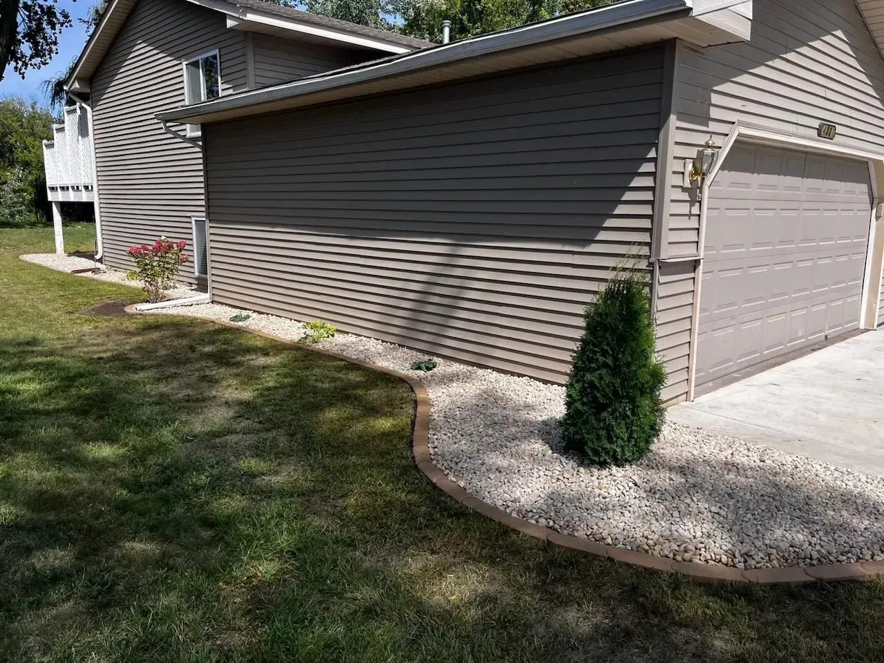 A house with gray siding and a two-car garage. A green shrub sits in a rock bed bordering the lawn.