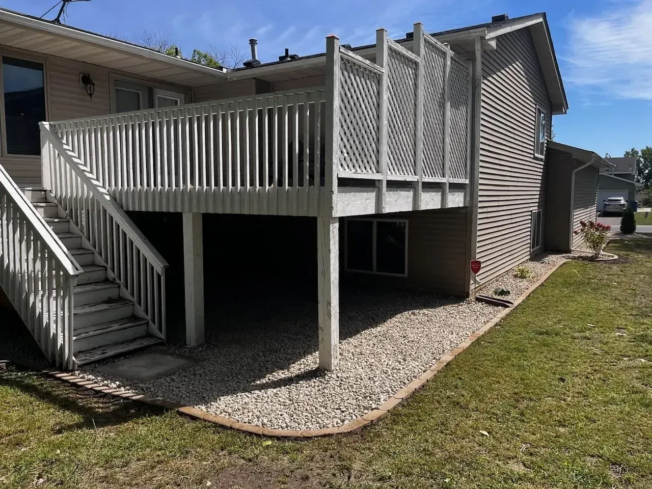Elevated wooden deck attached to a house with stairs, lattice panels, and gravel landscaping in the yard on a sunny day.