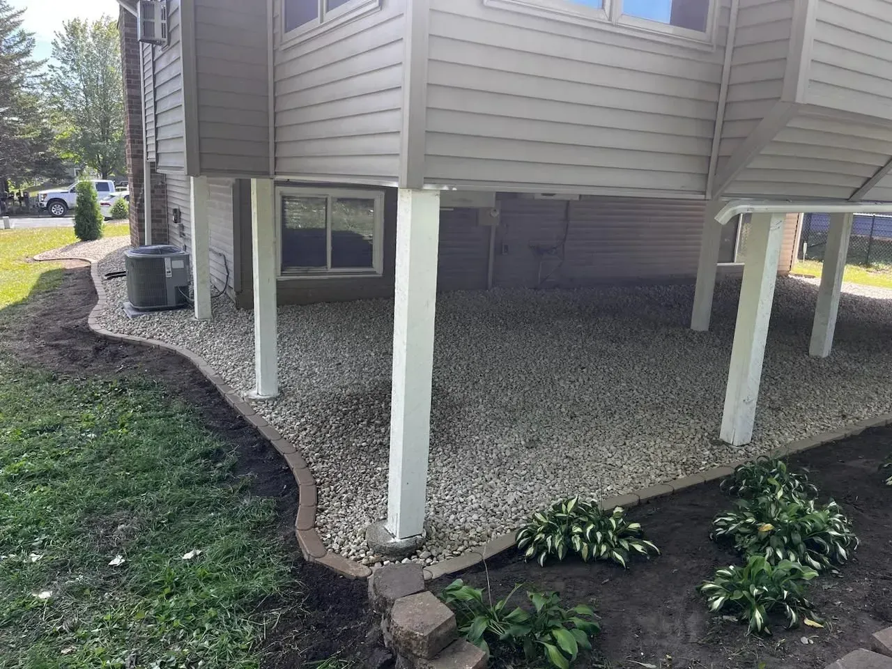 A gravel patio under an elevated house with beige siding, surrounded by a dark border and landscaping, including hostas.