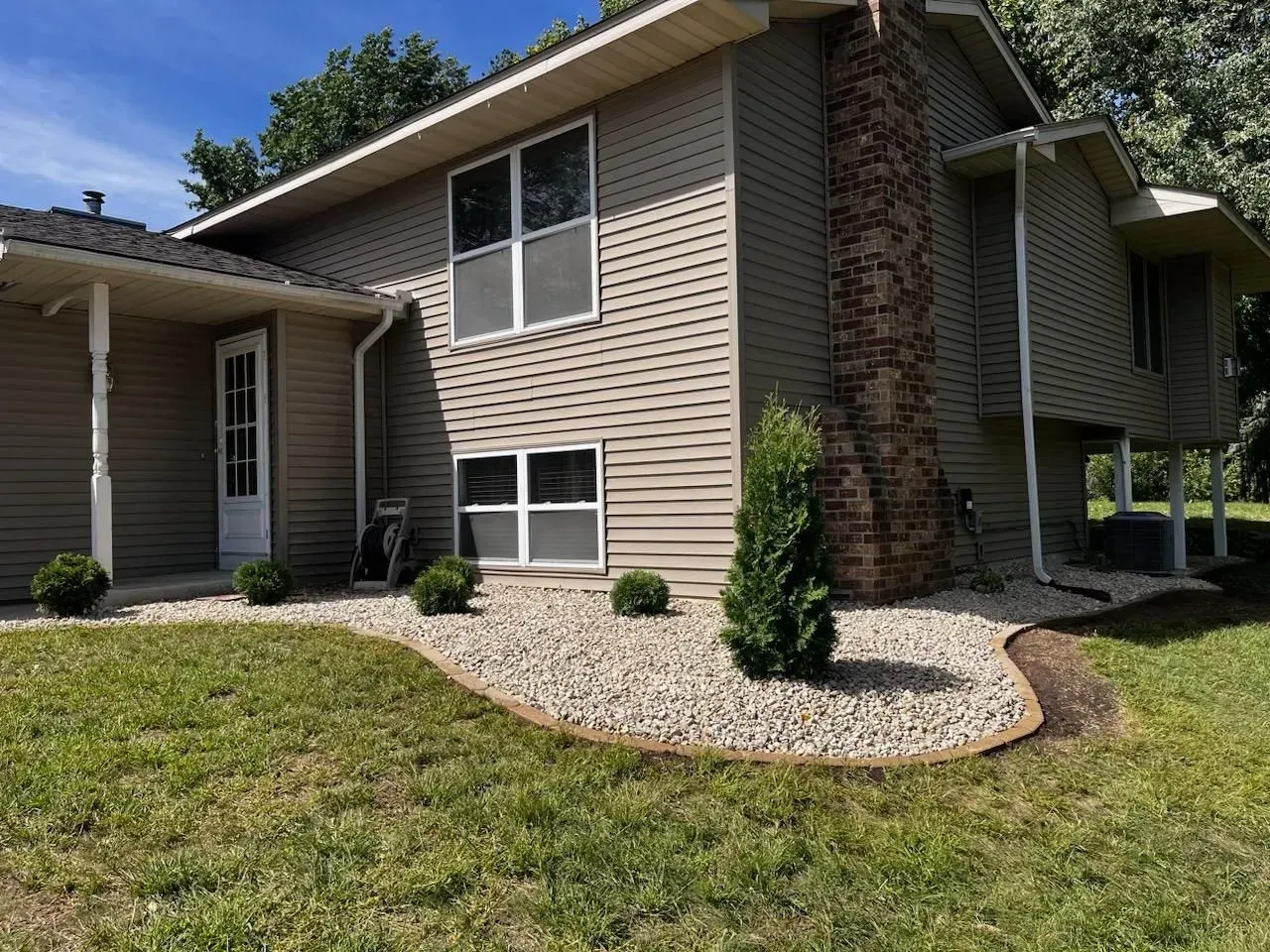 Beige house with stone chimney and landscaping. Gravel bed with green bushes borders the lawn.