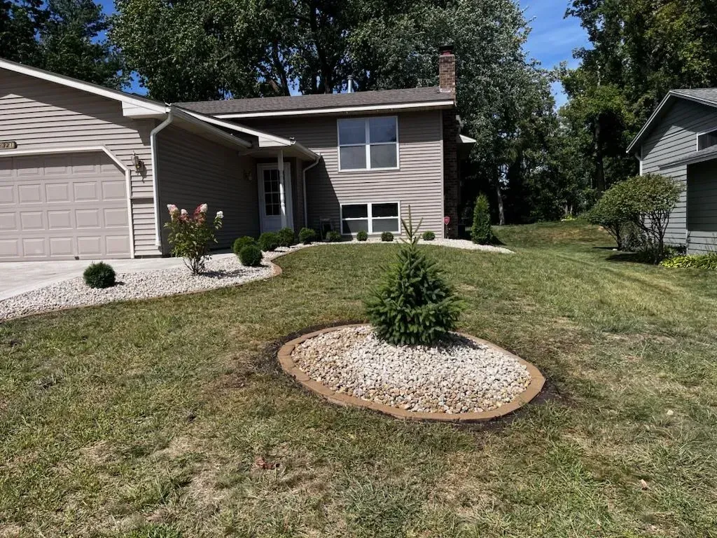 Tan house with a small evergreen tree in a rock bed in front. The yard has dry grass and other shrubs.