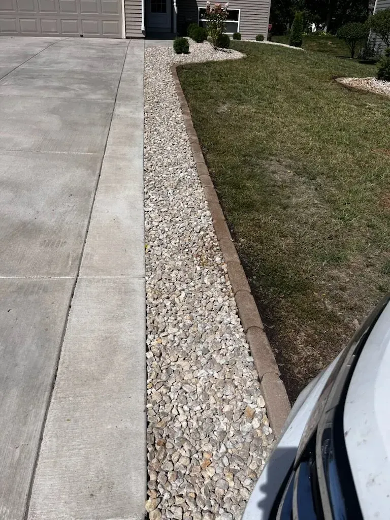 A driveway next to a gravel bed bordered by a concrete curb and lawn. A car's front is visible on the right.
