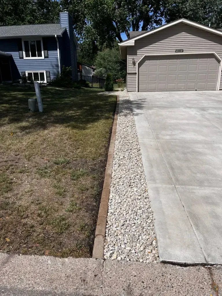 Driveway bordered by gravel, next to a grassy lawn and a blue house on the left, and a beige house and garage on the right.