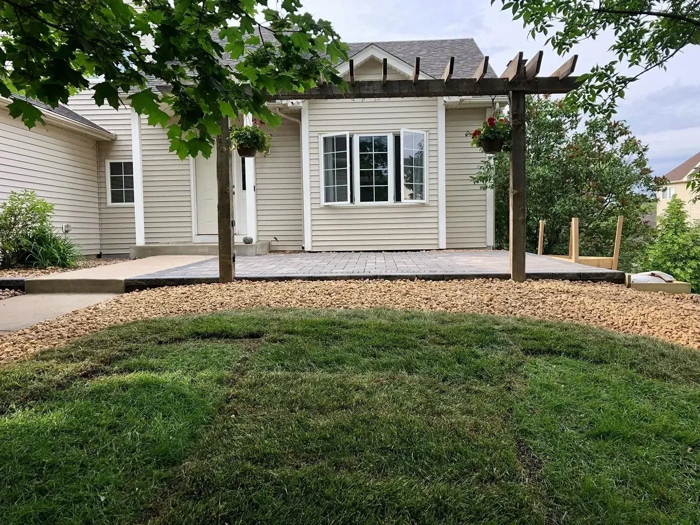 A beige house with a patio, pergola, and lawn. Two hanging flower baskets adorn the pergola. 