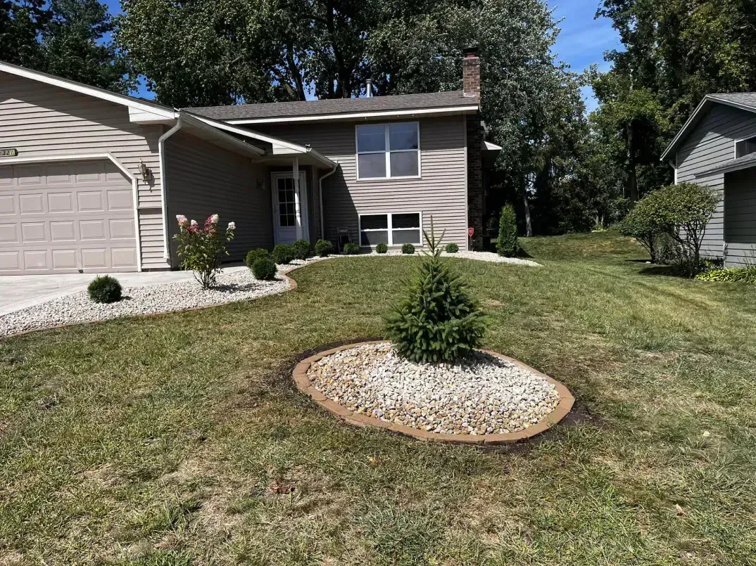 A house with a gravel bed surrounding a small evergreen tree in the front yard. Beige siding and green grass.