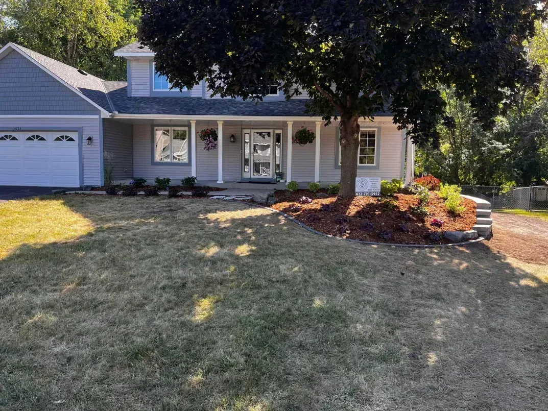 Gray house with white trim and a large tree shading the front yard. Brown mulch in the flower beds.