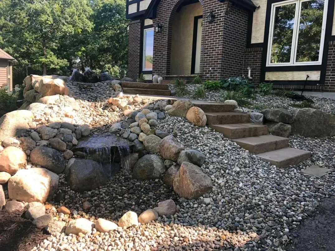 A tiered landscape with a small waterfall and stone steps leading to a brick house's entrance.