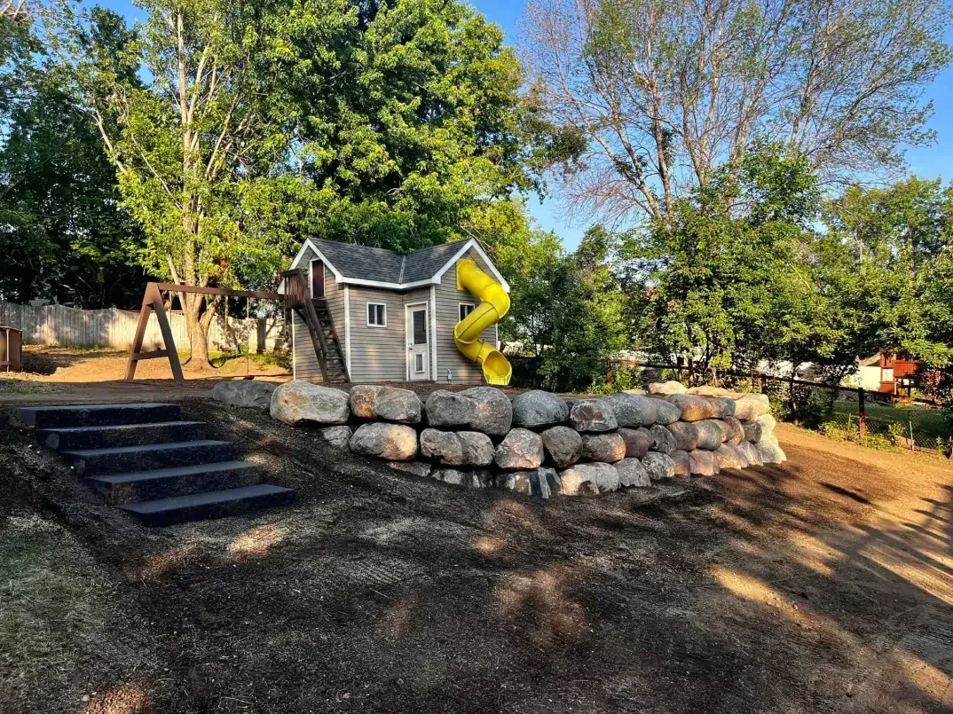 Playground with a small house, slide, and stone retaining wall. Steps lead up to the structure. Green trees and blue sky in 