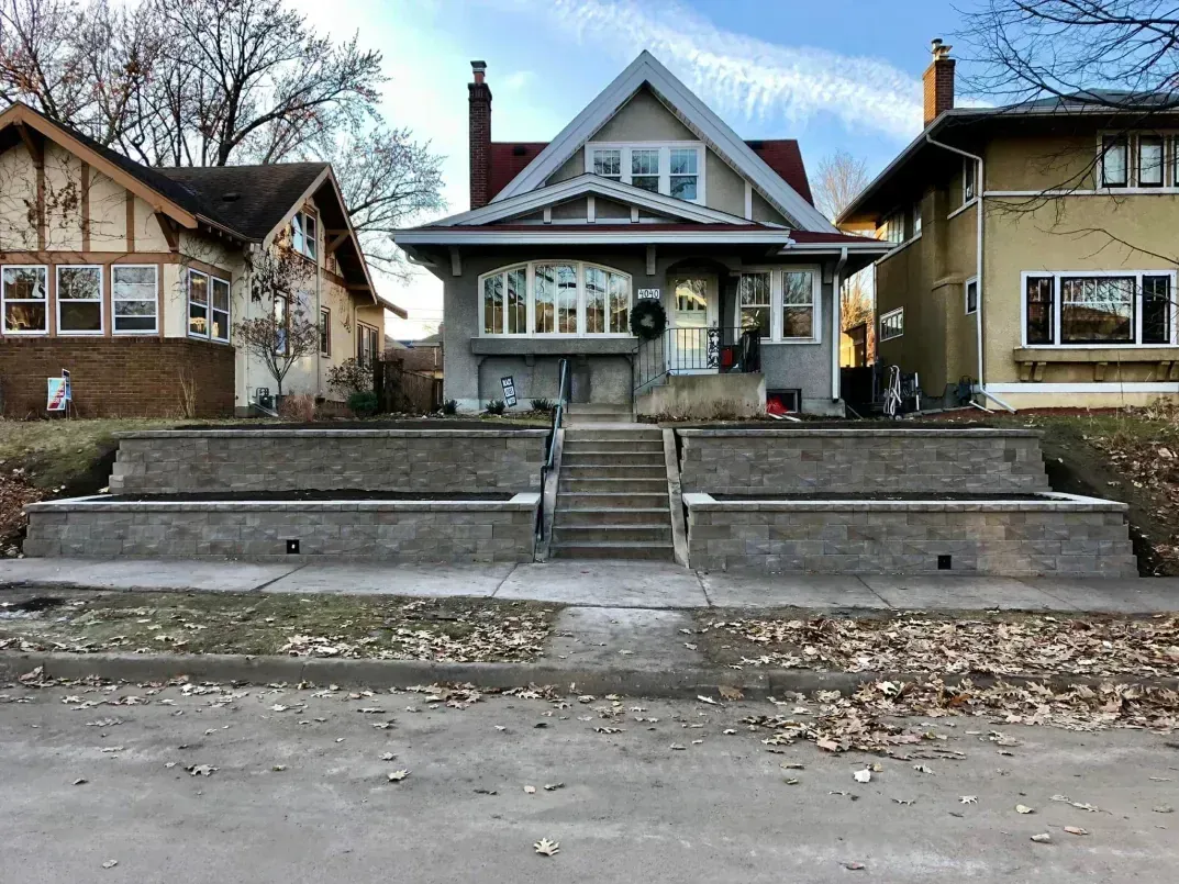 A gray two-story house with a front porch, set on a raised concrete foundation, flanked by other homes on a street.