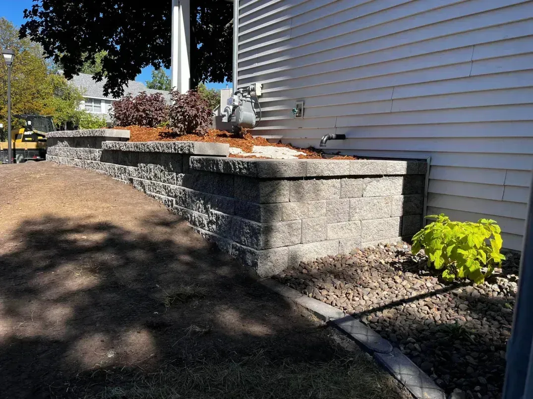 A brick retaining wall next to a house holds soil and landscaping with a gas meter visible.