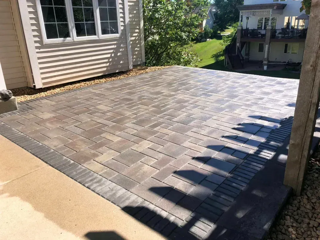 Brick paver patio next to a house with a beige foundation, bordered by a dark gray accent and small gravel.