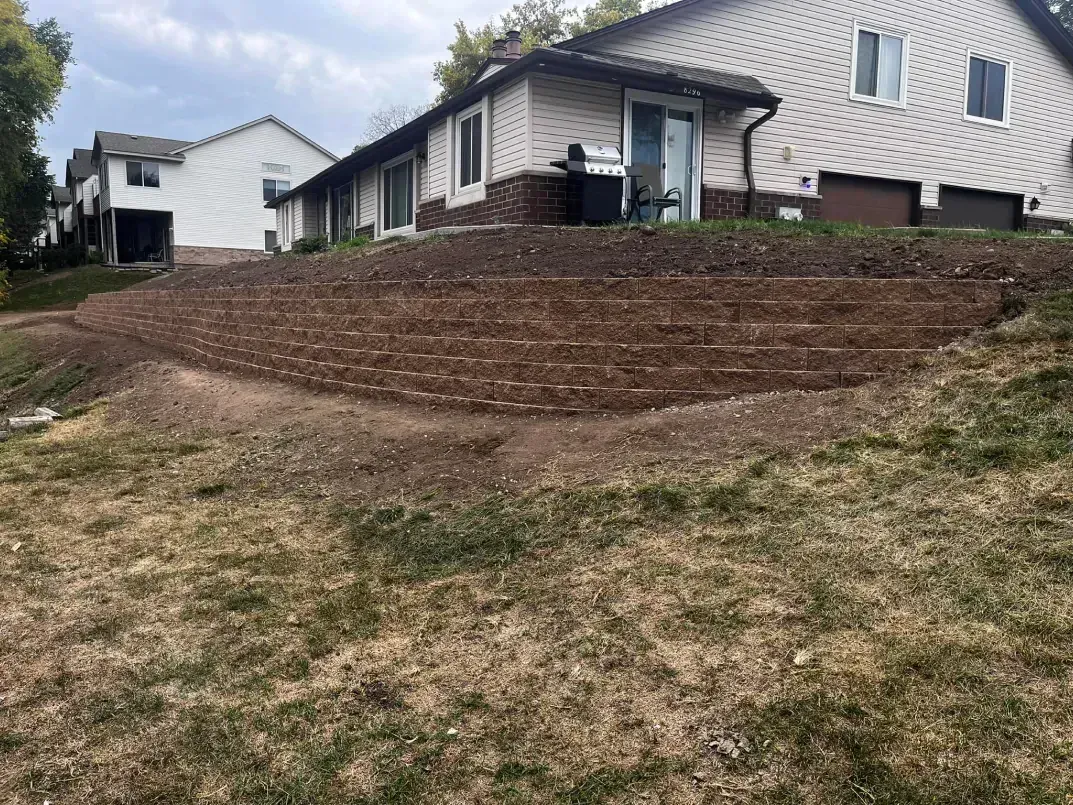 Graded hillside with newly planted soil in front of a suburban house. Brown soil, green grass, cloudy sky.