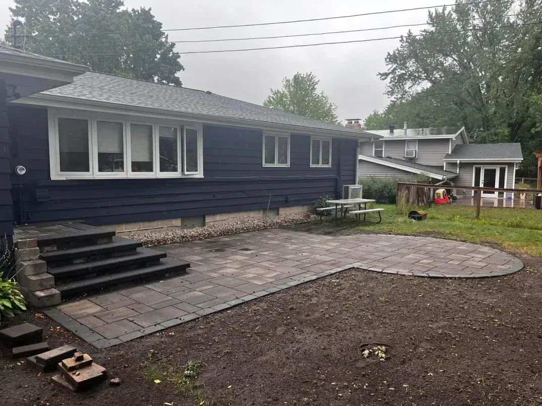 A newly constructed paved patio and steps next to a blue house. The patio curves outward into a yard with a picnic table.