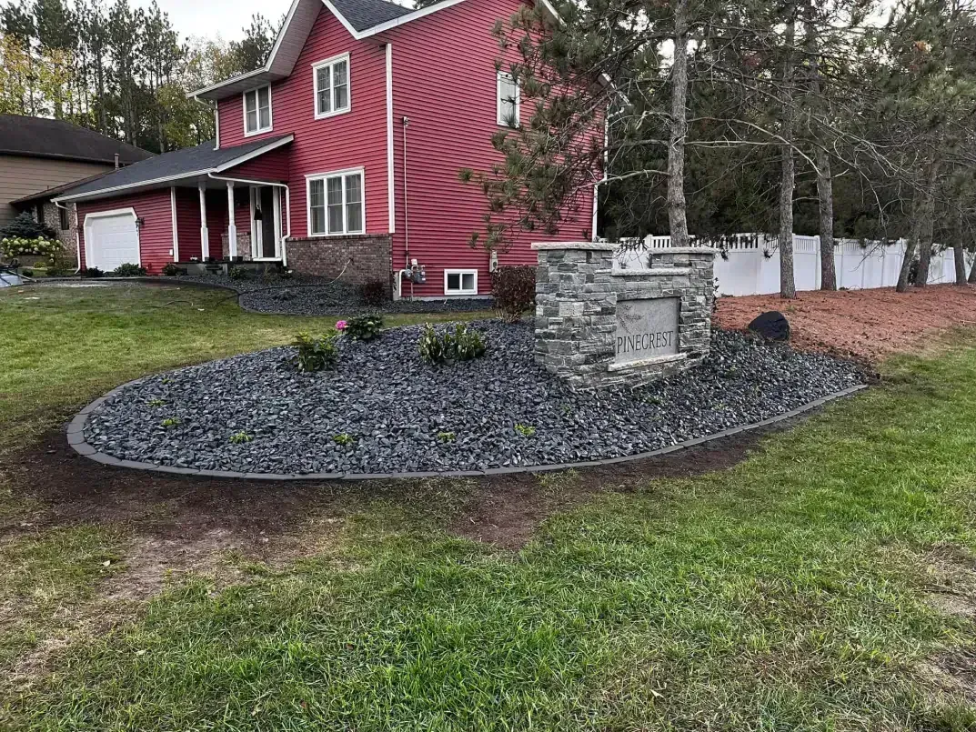 Red house with a stone monument in front, surrounded by black gravel and a curved border. Green lawn.