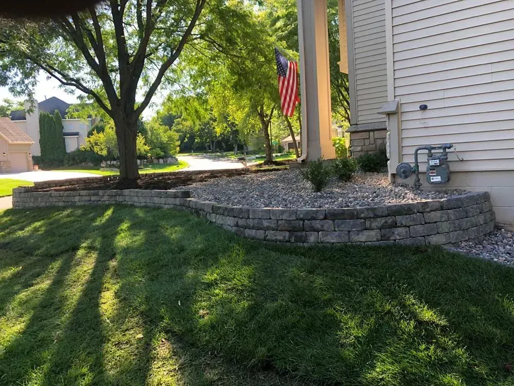 A landscaped front yard with a stone retaining wall, grass, and a tree near a house with an American flag.