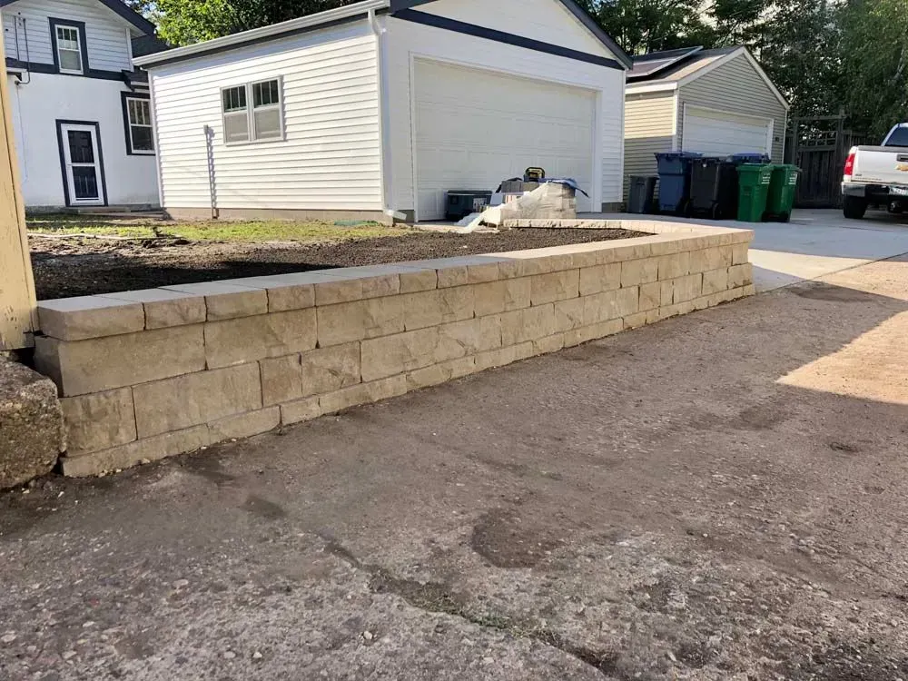 A beige retaining wall in a paved driveway, separating the pavement from a small patch of grass.