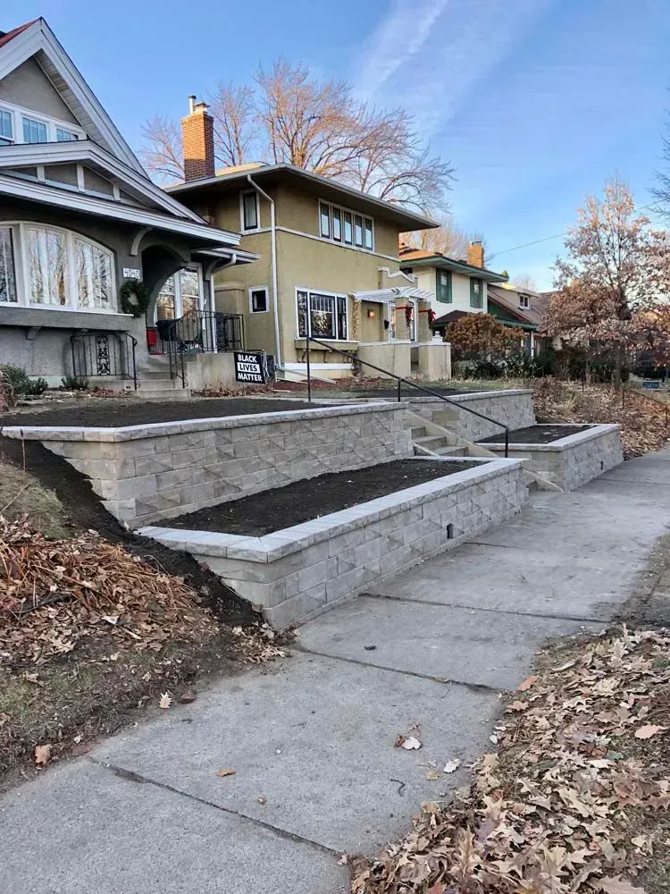 Stone tiered garden beds with black soil, a concrete pathway, and houses on a sunny day.