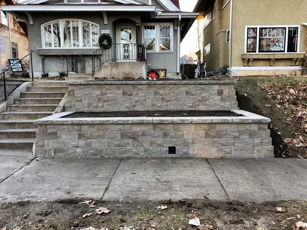 A front view of a house with stone retaining walls in front. Cement steps lead to the entrance. The sky is overcast.