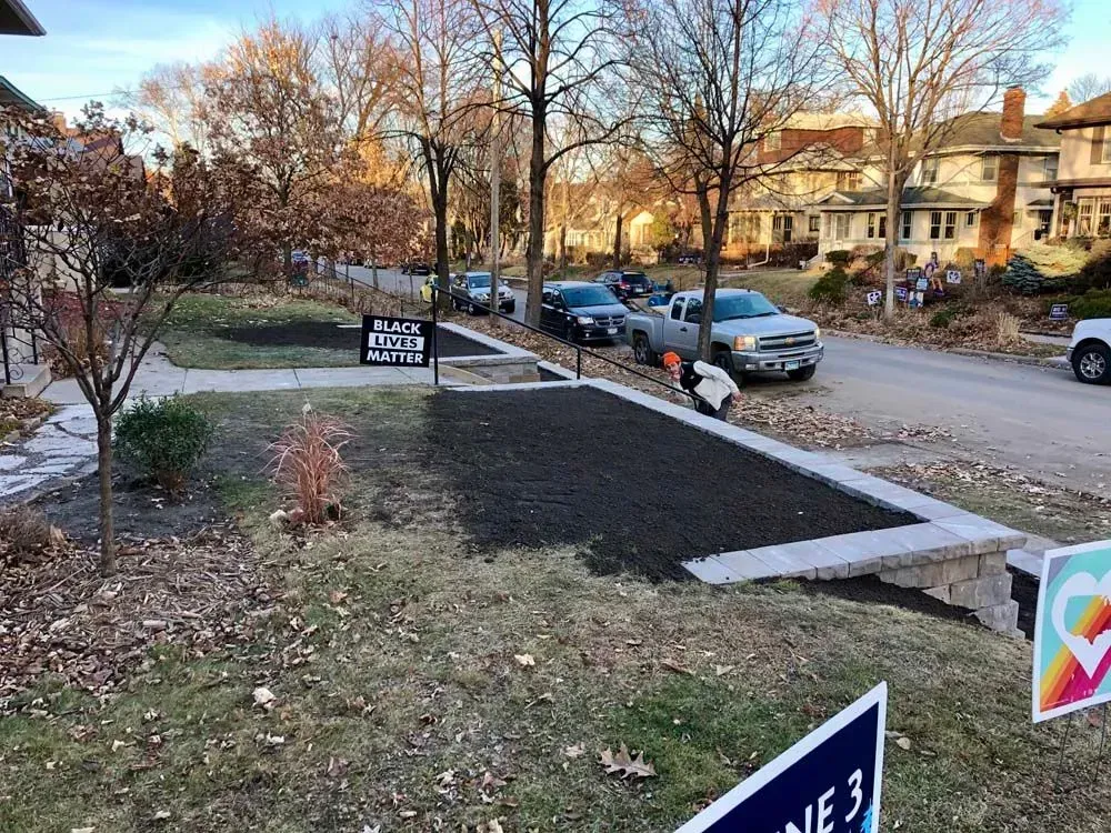 Residential street scene with cars parked along the curb, newly planted landscaping, and signs in yards.