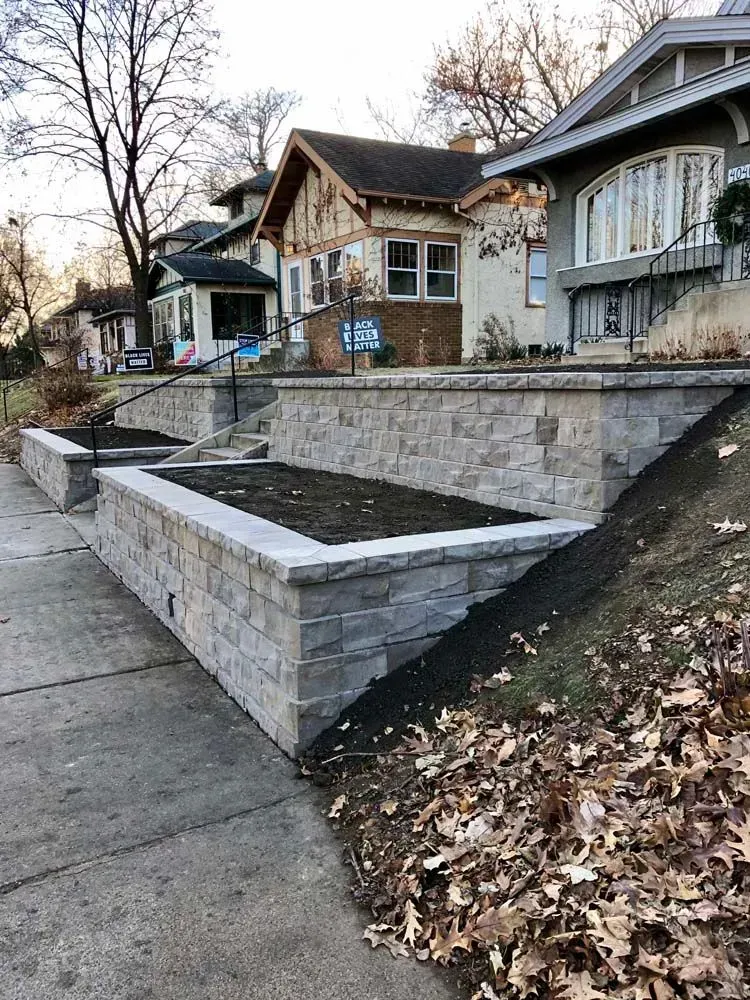Stone retaining walls with plants border a sloping residential street. Three houses are in the background.