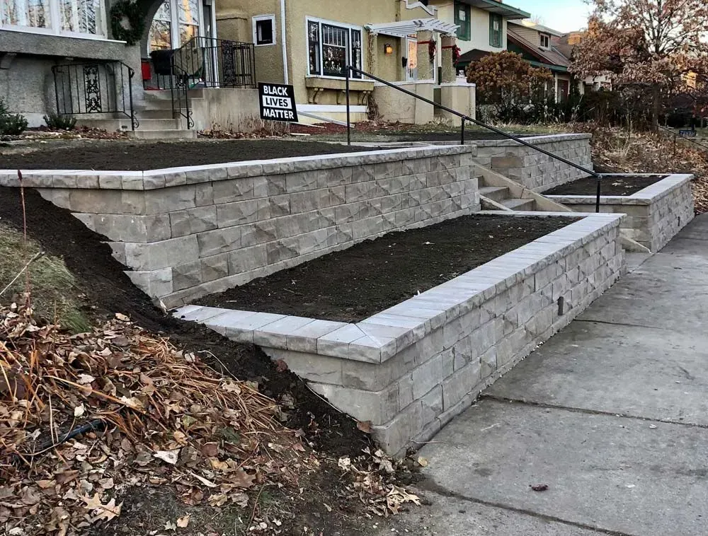 Stone retaining walls with built-in planters and stairs lead up to a house. Dark soil fills the planters.