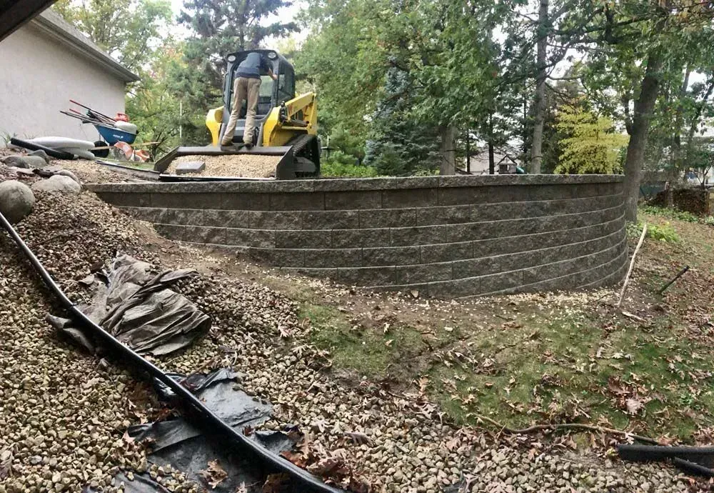 Construction of a curved retaining wall. A person operates a yellow skid steer. Earth and gravel surround the wall, with 