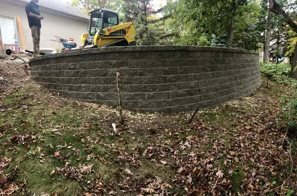 A curved retaining wall made of dark gray blocks being constructed outdoors, with a person and machinery visible.