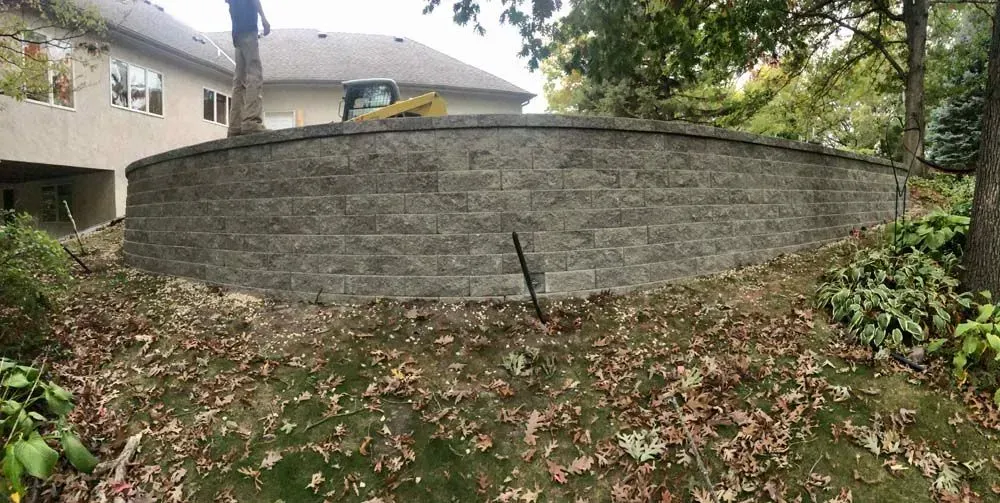 A curved retaining wall of gray stone blocks sits in a yard with fallen leaves and a house in the background.