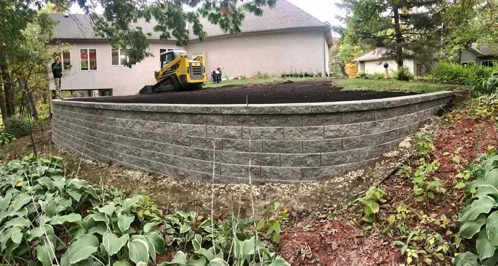 A retaining wall is built in a backyard with a small excavator on top, near a house.