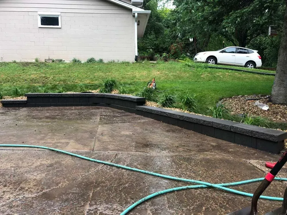 A concrete patio with a low retaining wall. A house, green grass, and a white car are in the background.