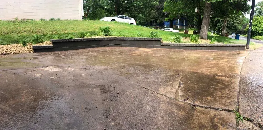 A wet concrete surface in front of a grassy hill, trees, and a house. The pavement shows cracks and a border wall.