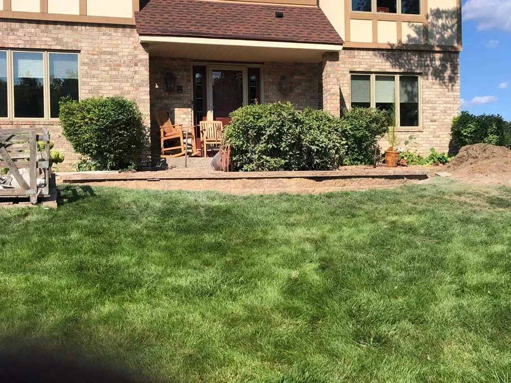 Backyard with a brick house, patio, & green lawn. Bushes frame the patio seating, & a small pile of dirt is off to the side.