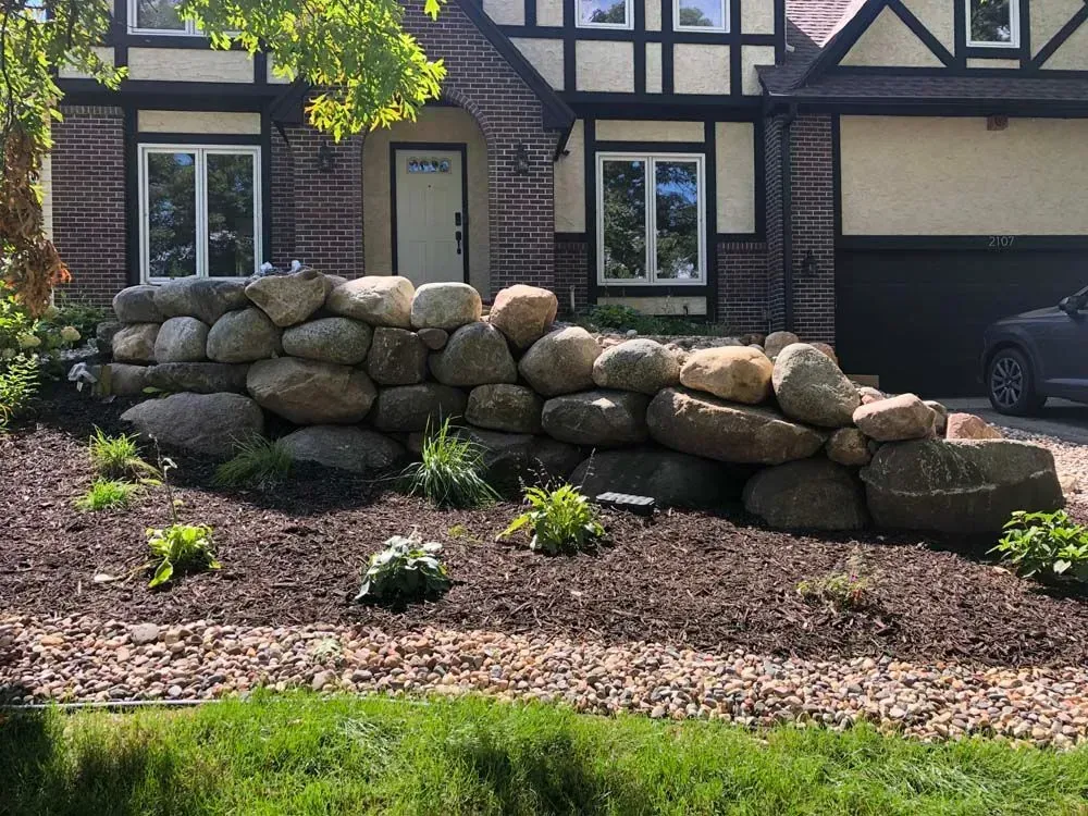 Stone retaining wall in front of a Tudor-style house, surrounded by mulch and greenery.
