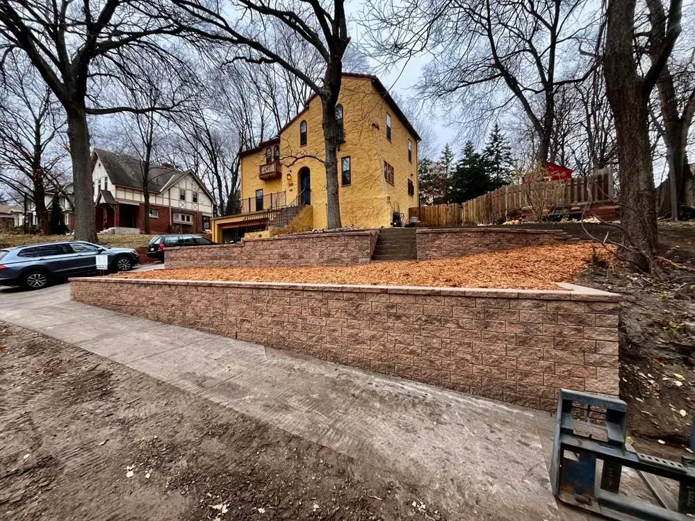A two-story yellow house with retaining walls and mulch landscaping. Trees and parked cars are visible in the setting.
