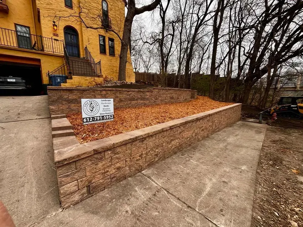 A tiered brick retaining wall with a sign, covered in fall leaves, beside a concrete driveway in front of a yellow house.
