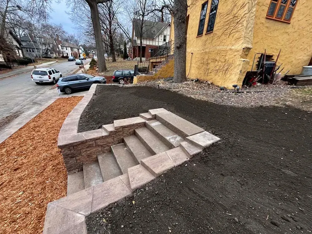 Stone steps and retaining wall lead up to a house on a residential street. Freshly tilled earth surrounds the steps.