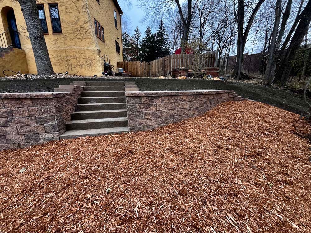 Brick retaining wall with stairs leading to a grass lawn in front of a yellow house.  Ground is covered in wood mulch.