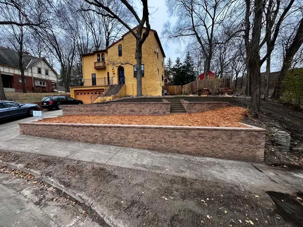 Yellow house with a tiered retaining wall in front, filled with brown mulch. Brown brick wall borders the sidewalk.