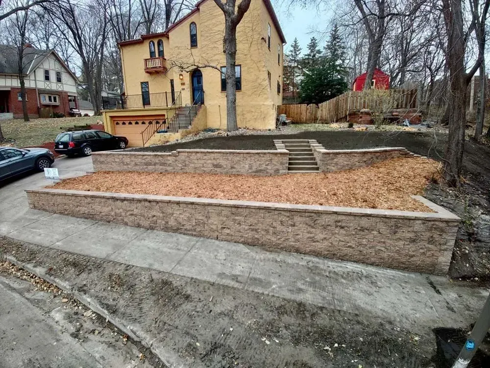 A tiered retaining wall with stairs leading up to a house on a sloped lot. Mulch covers the retaining wall surfaces.