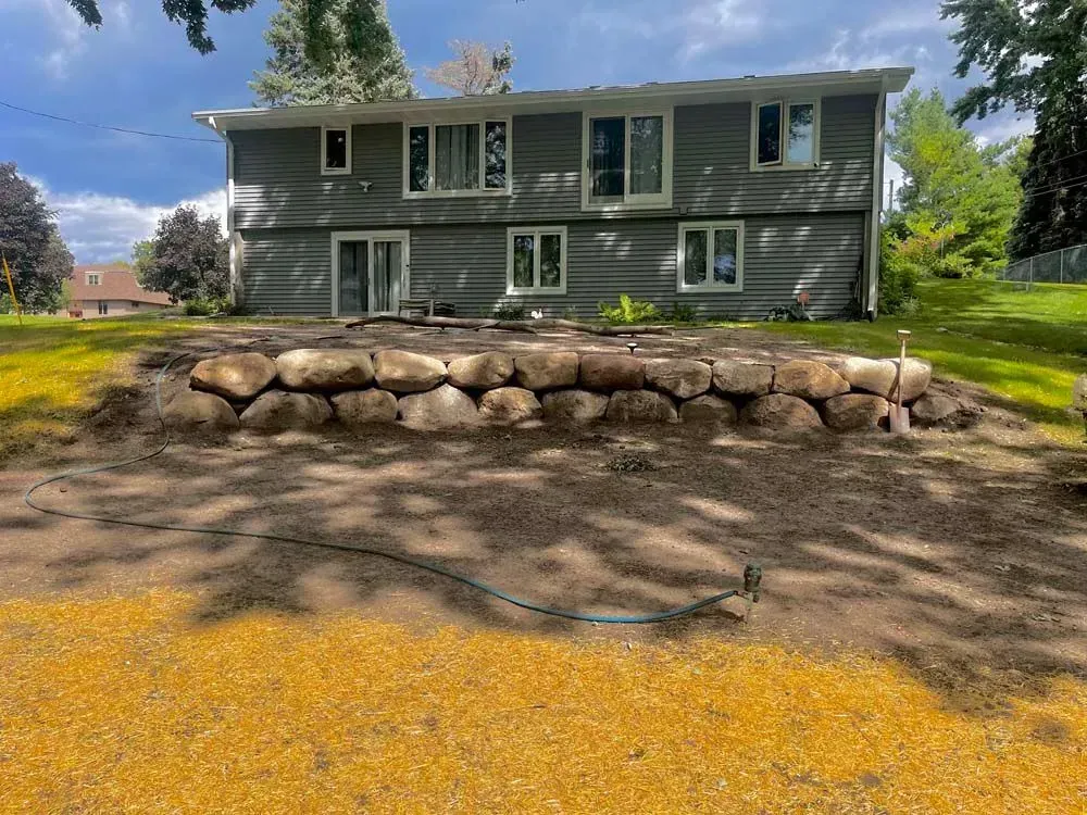 A two-story gray house with a stone retaining wall in the yard. A hose lies on the foreground dirt.