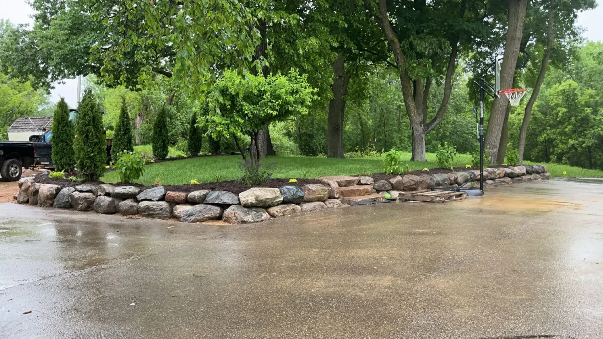 A wet driveway with a stone retaining wall with landscaping. Green trees and a basketball hoop are in the background.