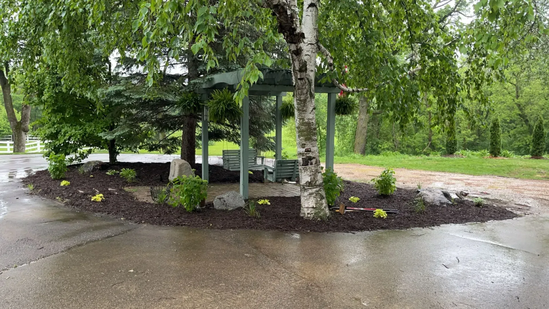 A gazebo with seating nestled under trees, surrounded by dark mulch and new plantings, on a wet asphalt surface.