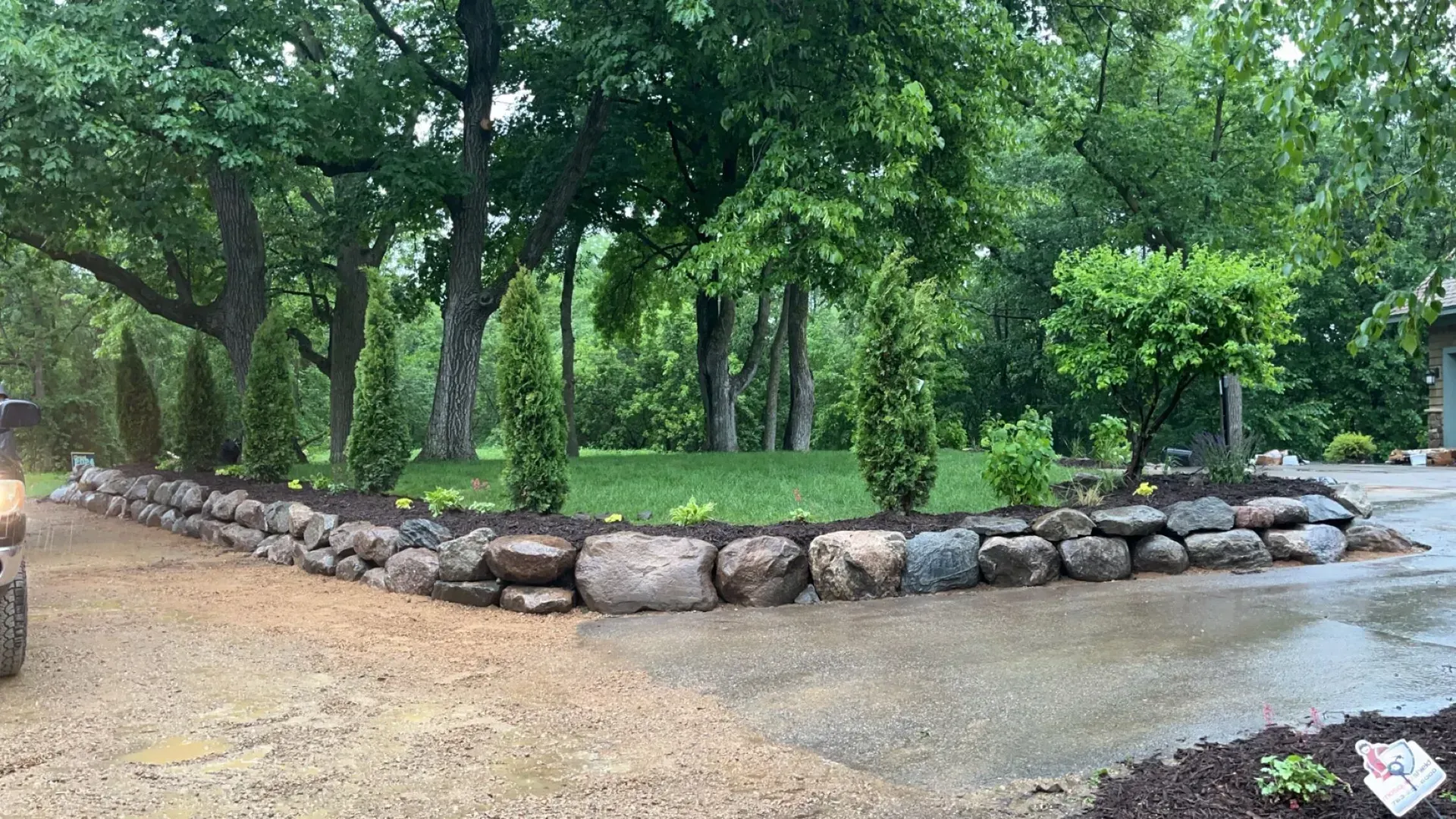 Stone retaining wall with trees and freshly planted mulch. A driveway leads toward a green lawn and wooded area.