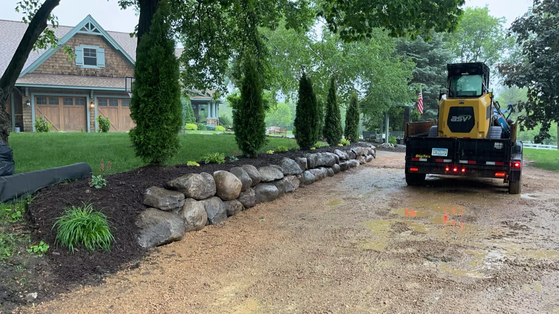 A large rock wall lines a muddy driveway, with landscaping and a trailer carrying construction equipment. 