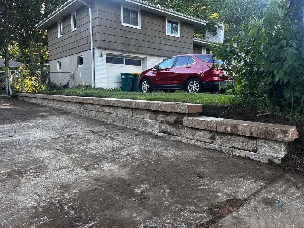 Stone retaining wall next to a driveway, a red car parked in front of a two-story house.