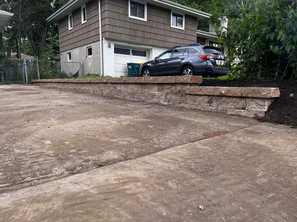 Concrete driveway with a low retaining wall in front of a two-story house with a car parked in the driveway.